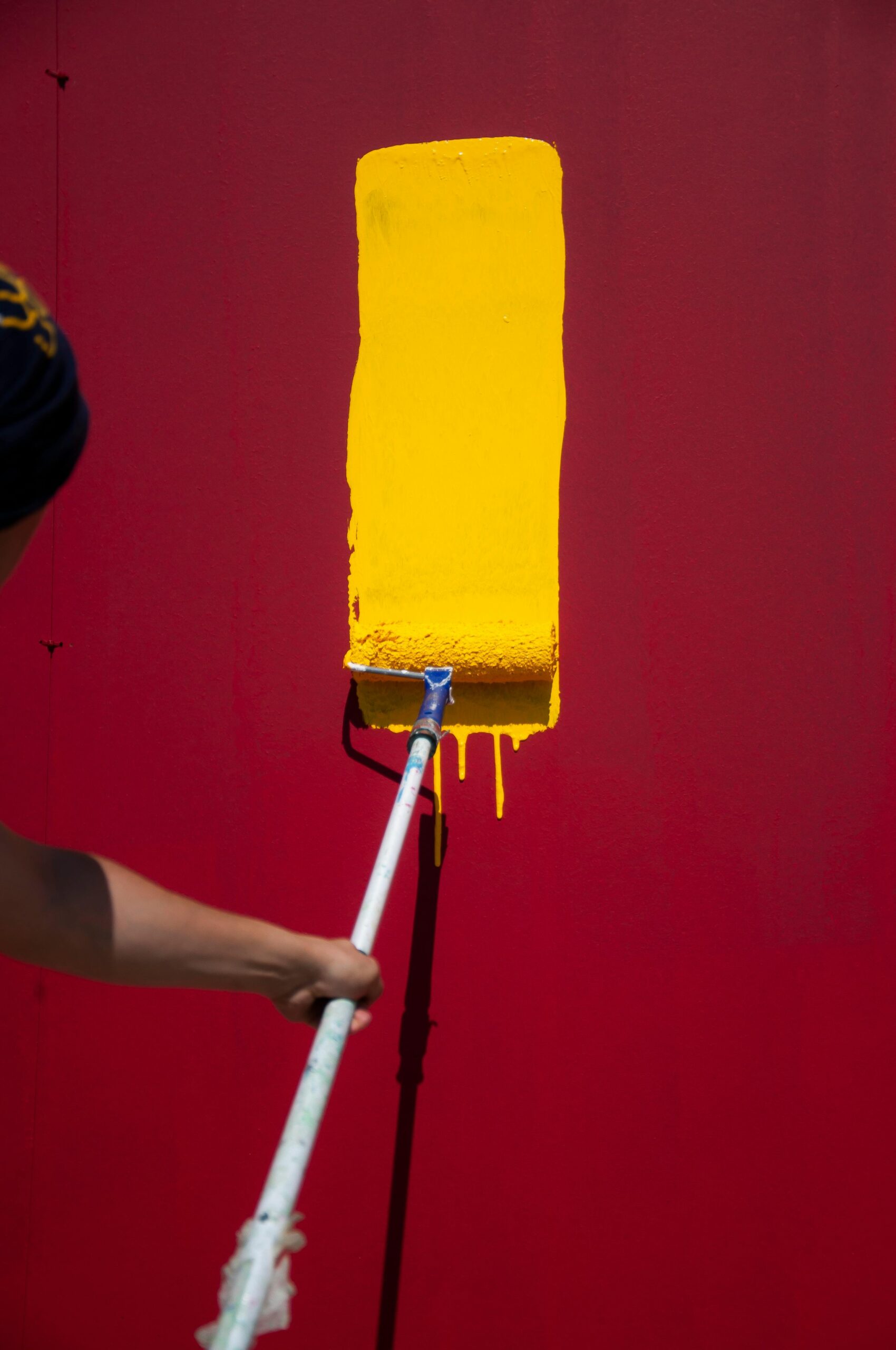 Close-up of a person using a roller to paint a yellow stripe on a red wall.